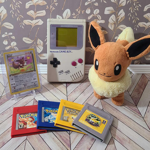 A plush Eevee sits beside a Game Boy, four classic Pokémon game cartridges, and an Eevee trading card