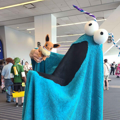 A person in a bright blue Yip Yip Martian costume with big white eyes and twisted antennae holds an Eevee plush at Tekko 2025, with other cosplayers visible in the background